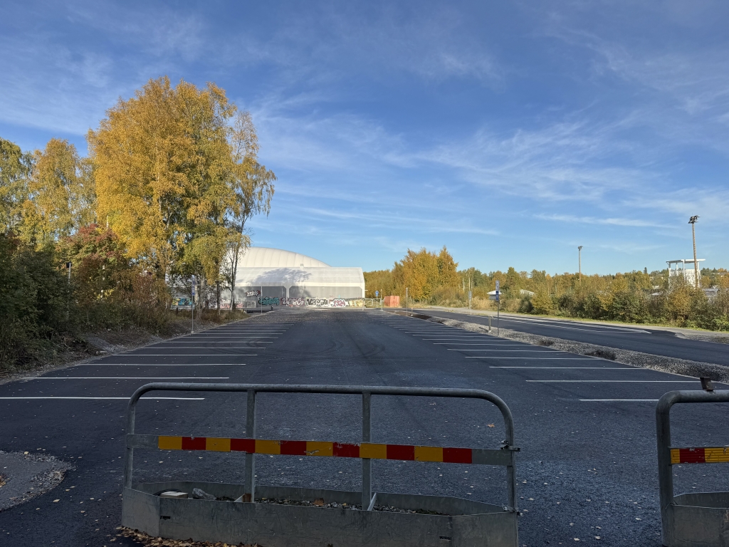 Asfaltoitu pysäköintialue, jossa on merkittyjä pysäköintiruutuja. Alueen etuosassa on puomit, ja taustalla näkyy puita sekä suuri hallirakennus.