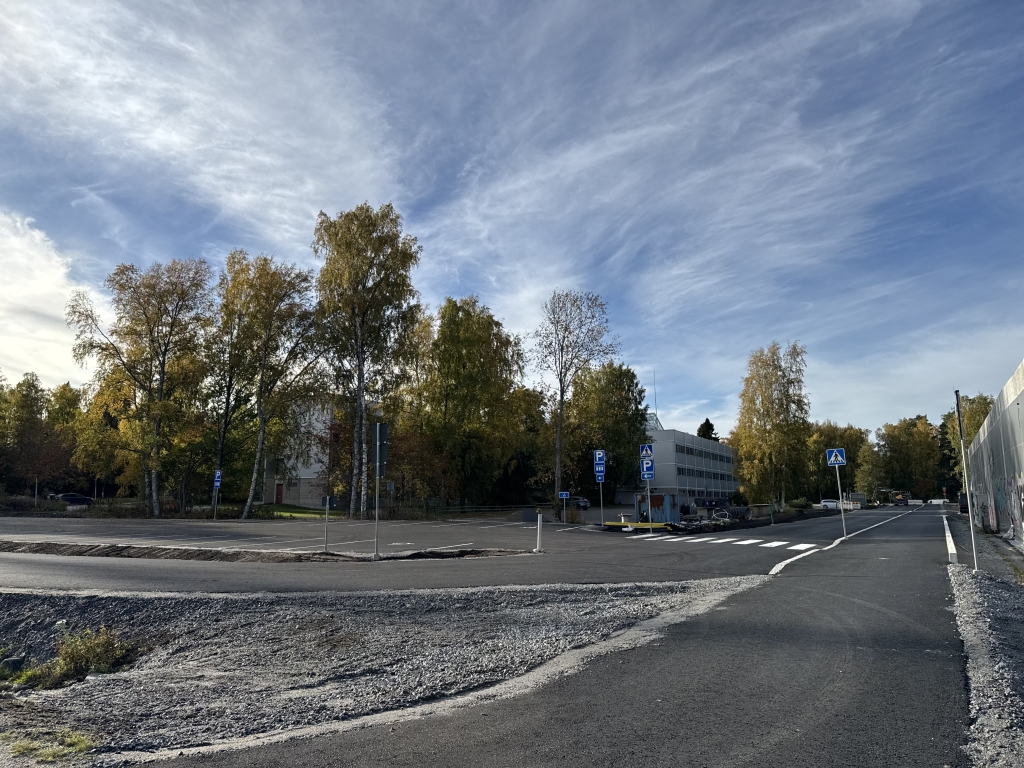 Asfaltoitu katu ja pysäköintialueen liittymä, joiden yhteydessä on suojateitä, liikennemerkkejä ja pysäköintialueen opasteita. Aluetta ympäröivät puut ja rakennukset.