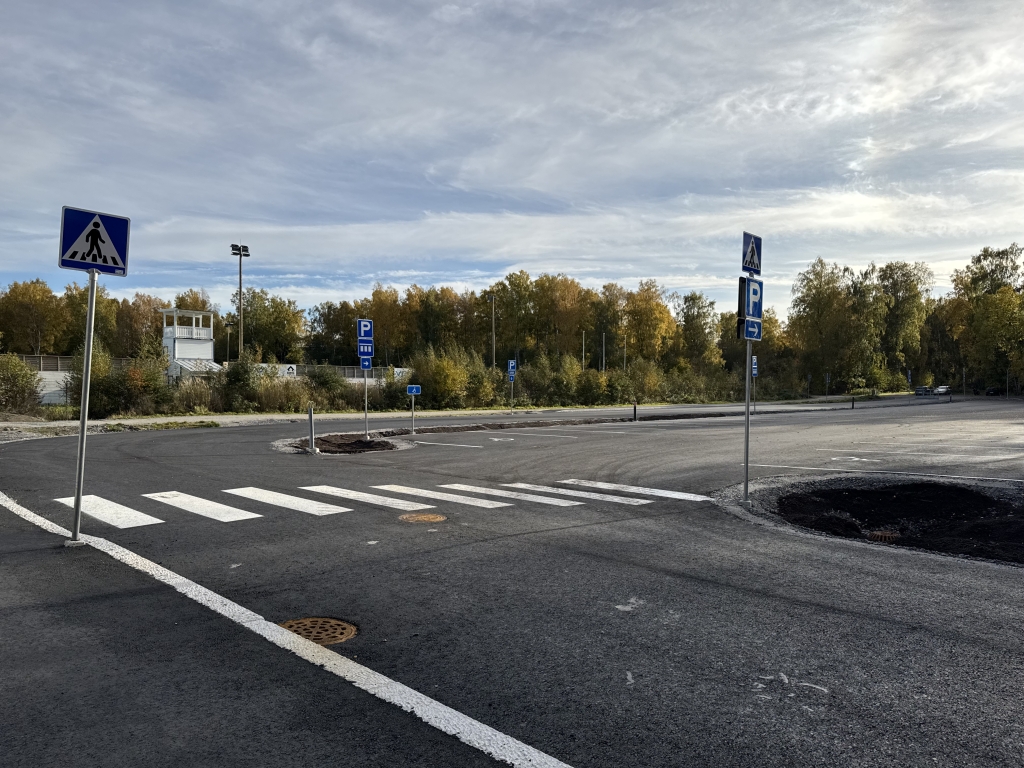Asfaltoitu katu ja pysäköintialueen liittymä, joiden yhteydessä on suojateitä, liikennemerkkejä ja pysäköintialueen opasteita. Aluetta ympäröivät puut ja rakennukset.