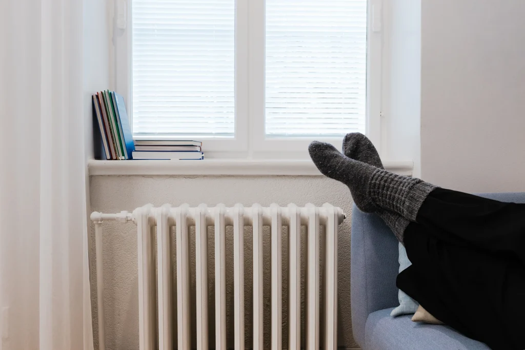 Henkilö rentoutuu sohvalla lämpöpatterin vieressä. Ikkunasta tuleva luonnonvalo ja lämmin sisätila havainnollistavat kodin lämmitystä ja asumismukavuutta.