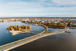 Flygfoto över staden vid havet. I förgrunden finns en terrasserad väg och en liten ö, i bakgrunden en stad med höga byggnader. Himlen är blå och i träden höstens färger.