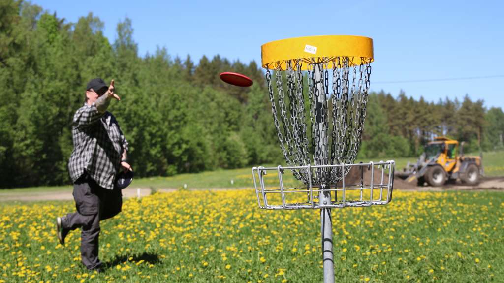 Mies heittää frisbeetä koriin, taustalla ajaa pyöräkuormaaja maata