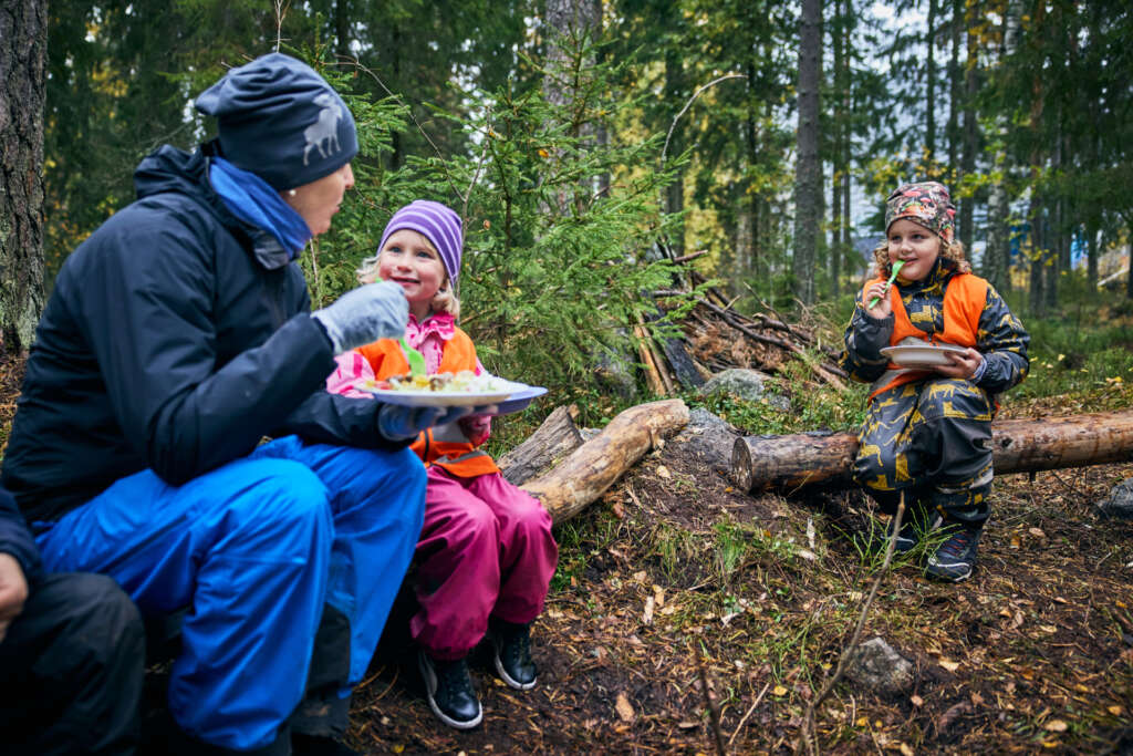 Aikuinen ja kaksi lasta istuvat metsässä kaatuneen puu päällä ja syövät lounasta.