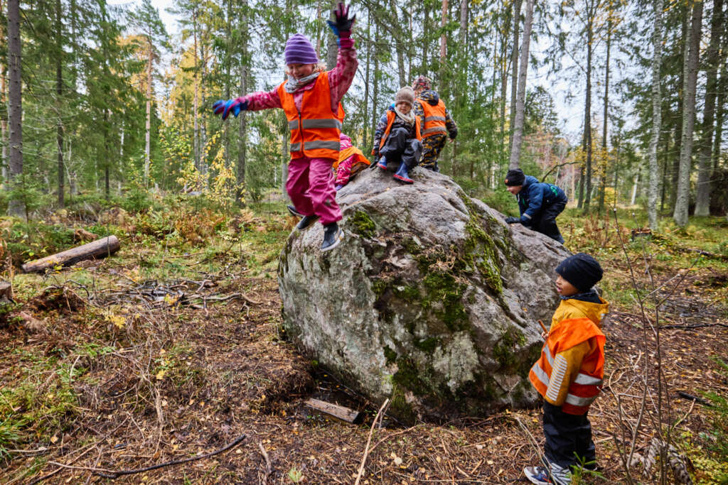 Lapset hyppivät ja kiipeilevät isolla kivellä metsässä.