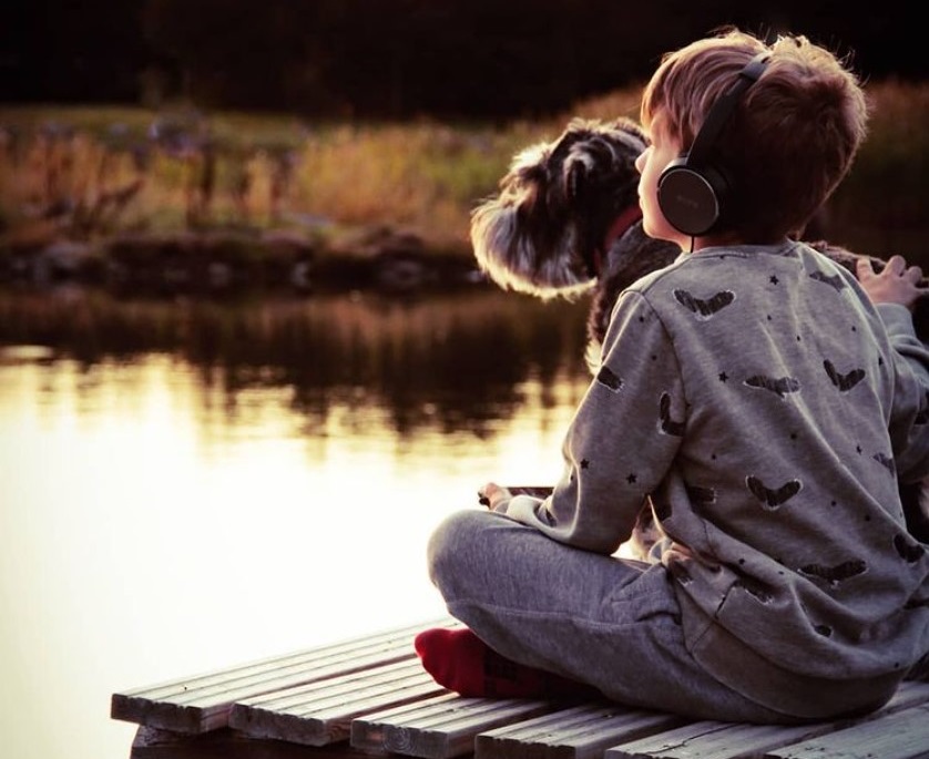 A boy sitting on a pier with a dog. The boy has headphones.
