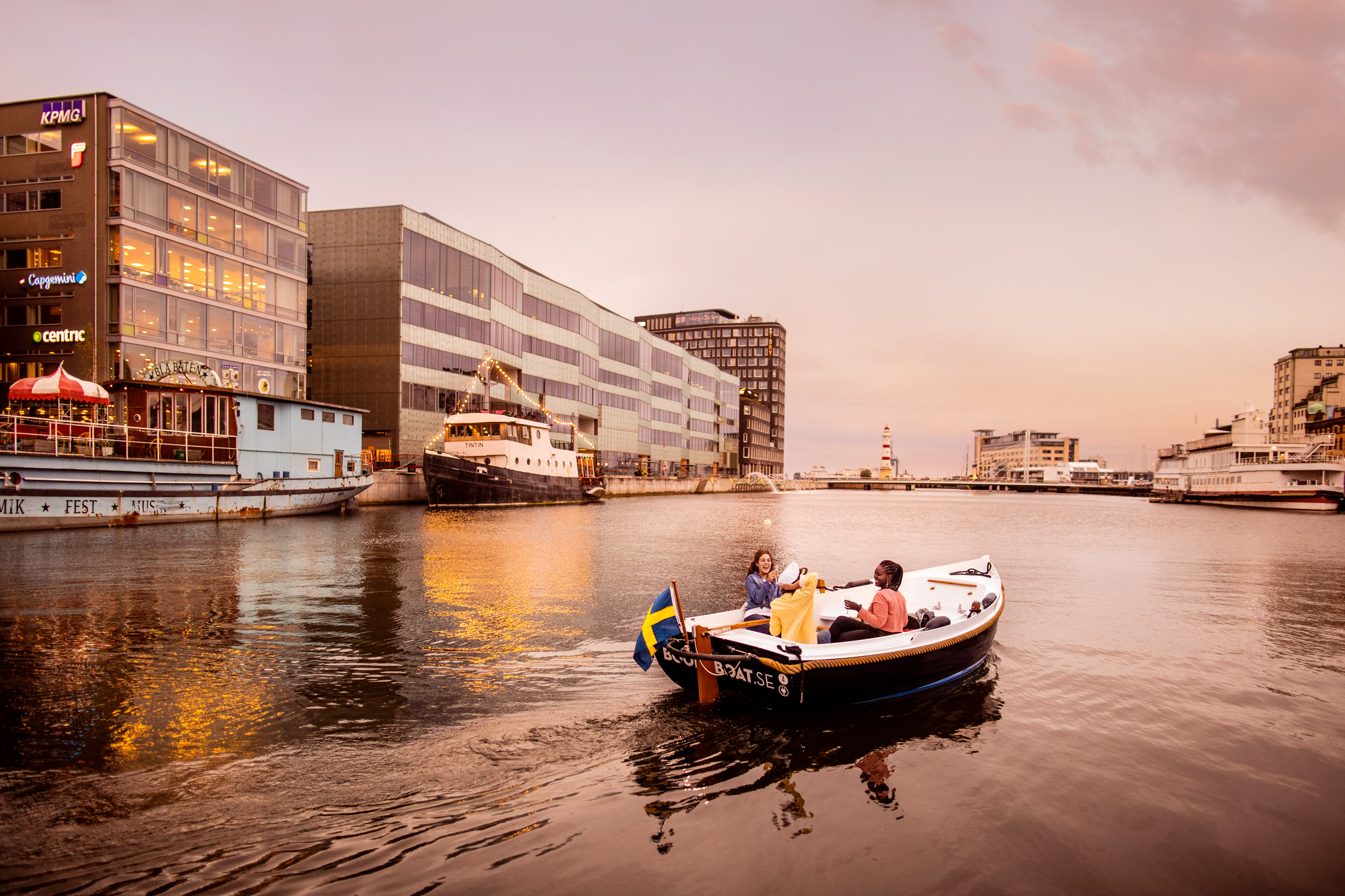 Kolme ihmistä veneilee Malmön kanavalla, taustalla Malmön yliopisto.