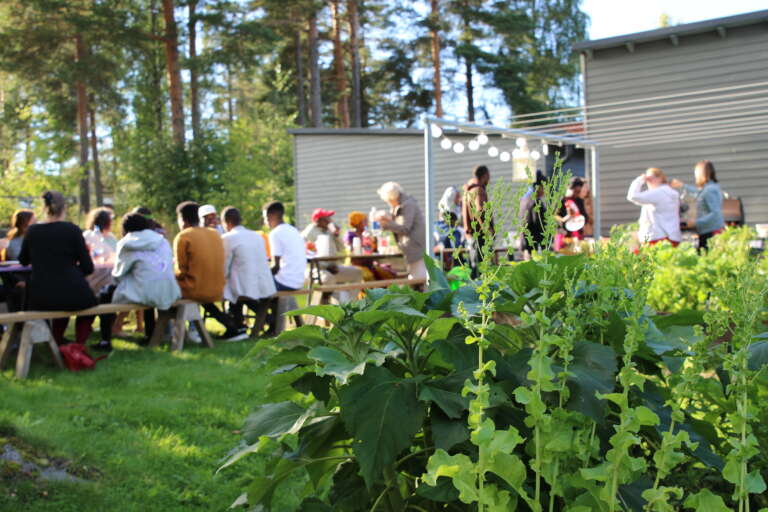 I främre delen av bilden ser man grönsaker som växer i en odlingslåda, i bakgrunden sitter en grupp människor vid ett långt trädgårdsbord.