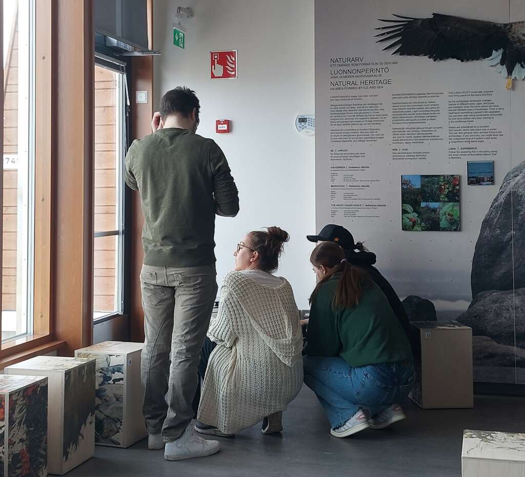 Neljä henkilöä tutkimassa esineitä näyttelytilassa.