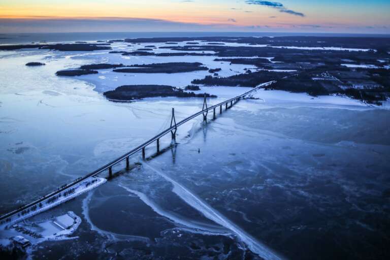 A long bridge from a bird's view, surrounded by frozen sea and islands in the horisont.