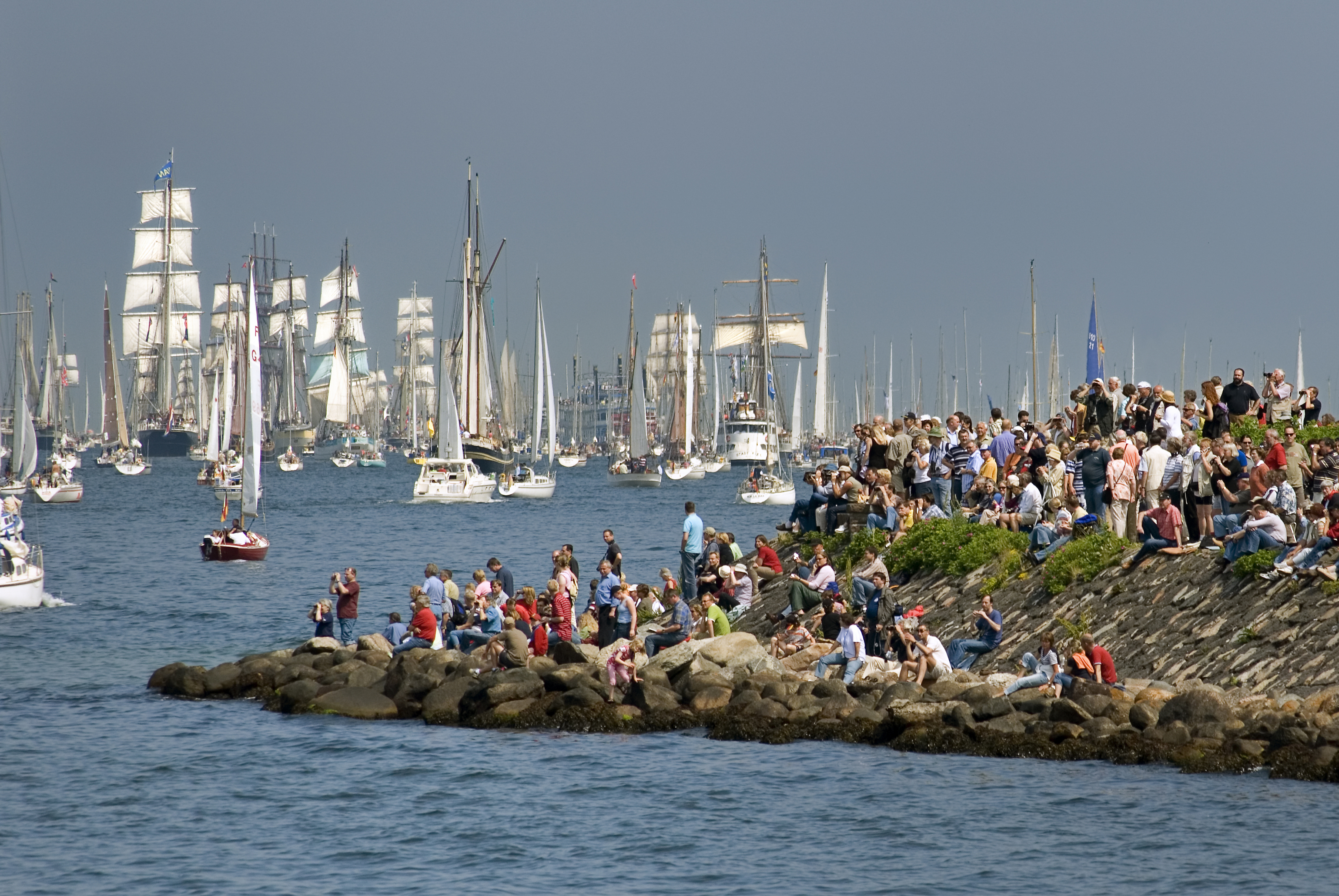 People sitting at the shore and watching hundreds of sailing boats.