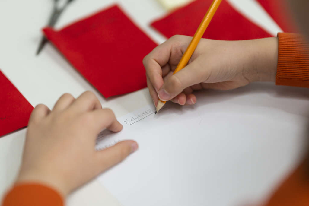 Lähikuva lyijykynällä kirjoittavan lapsen käsistä pöydän ääressä. Lapsi on kirjoittanut valkoisele paperille "Kehitettävää", pöydällä näkyy lisäksi sakset ja punaisia kartonkeja.