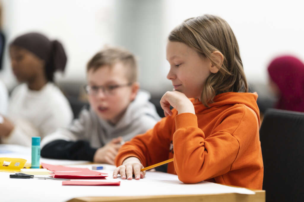 Närbild på en leende pojke i klass 5 som sitter vid ett bord. Framför sig har han pysselmaterial. Bakom honom kan man otydligt se två andra barn vid ett bord.