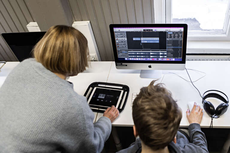 Oppilas editoi tietokoneella äänitiedostoa. Opettaja on vieressä ja hänella on ipad kädessä.