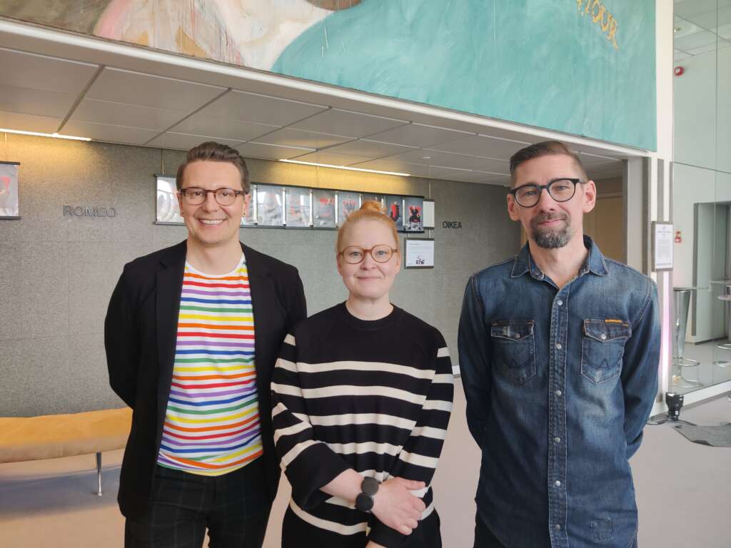 Kuvassa kolme henkilöä seisovat teatterin aulatiloissa ja katsovat kameraan.
