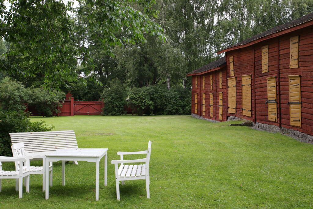 Vanhanaikaisia tuoleja ja pöytiä nurmikolla, taustalla puita ja punainen puutalo.