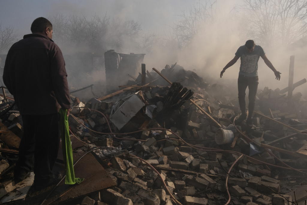 Two men walk among the ruins of their house. Dust hovers over the ruins. One man holds a green cloth in his hand.