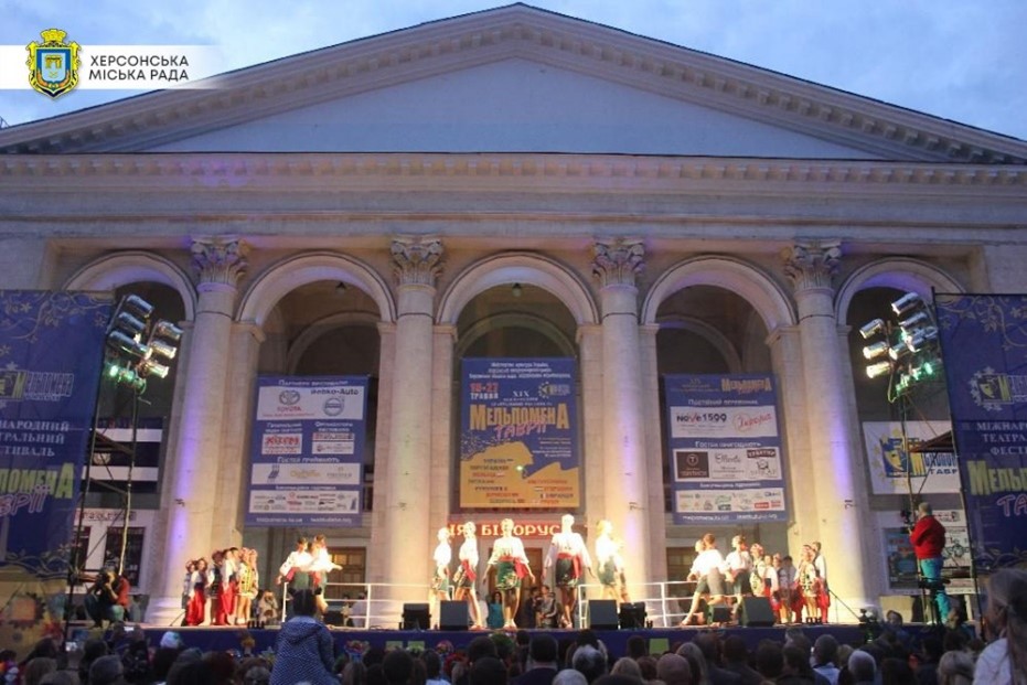 A stage in front of a Greek-style building with columns. People are performing in traditional clothes on the stage in front of an audience.