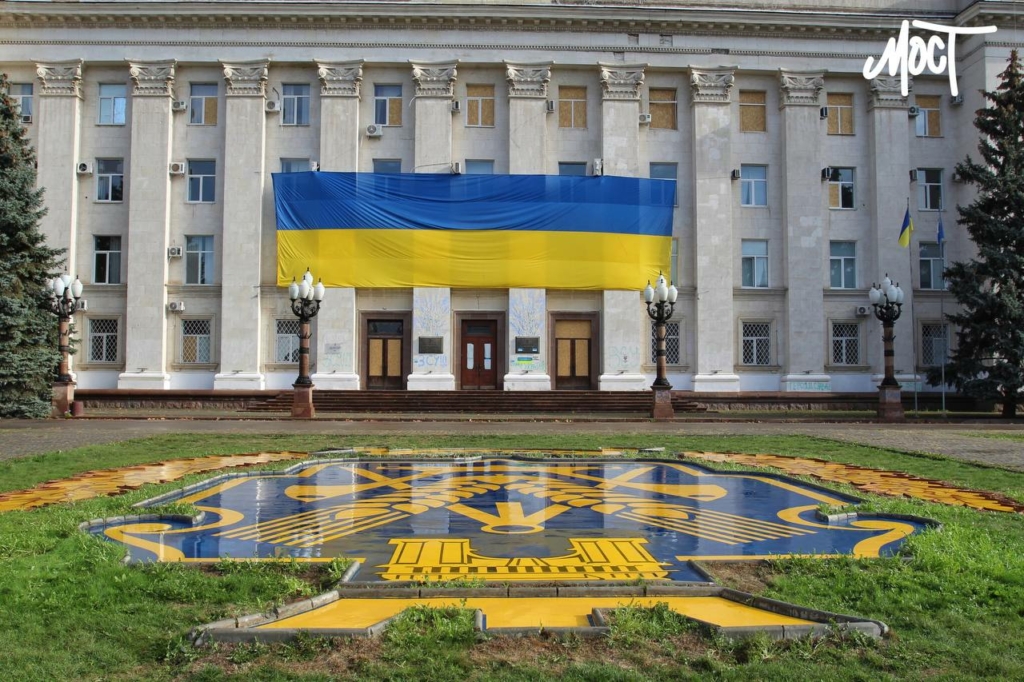 Chersons stadshus med en ukrainsk flagga på fasaden och stadens emblem på marken framför byggnaden.