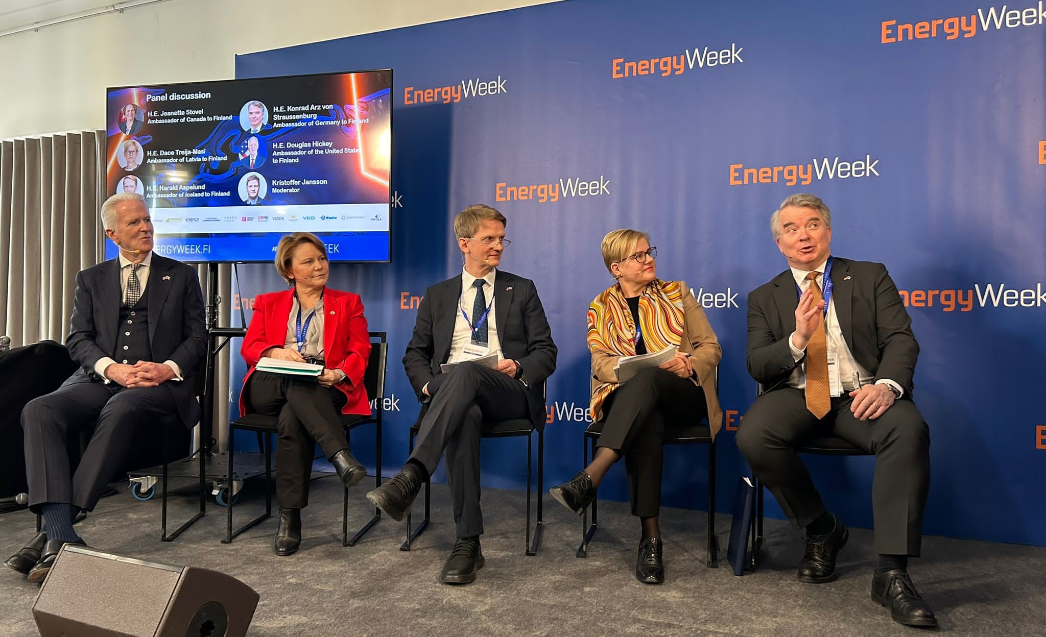 Five people sitting on chairs on a stage. In the background a wall with EnergyWeek on it.