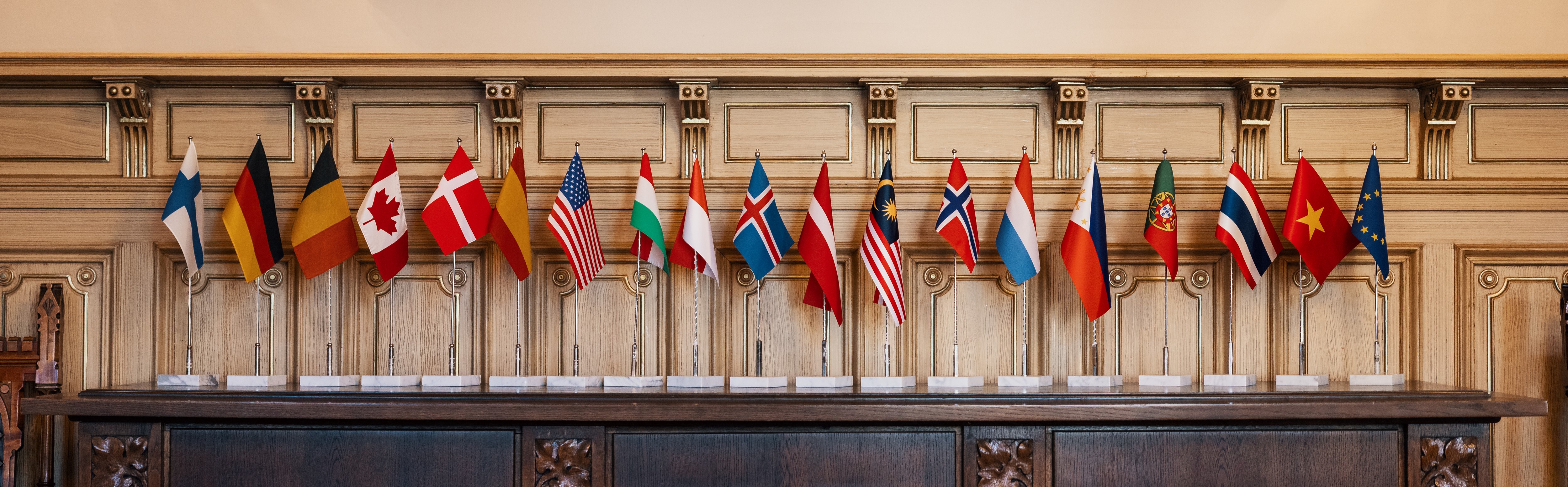 Several country flags on a table. From left to right: Finland, Germany, Belgium, Canada, Denmark, Spain, USA, Hungary, Indonesia, Iceland, Latvia, Malaysia, Norway, Netherlands, Philippines, Portugal, Thailand, Vietnam, and EU.
